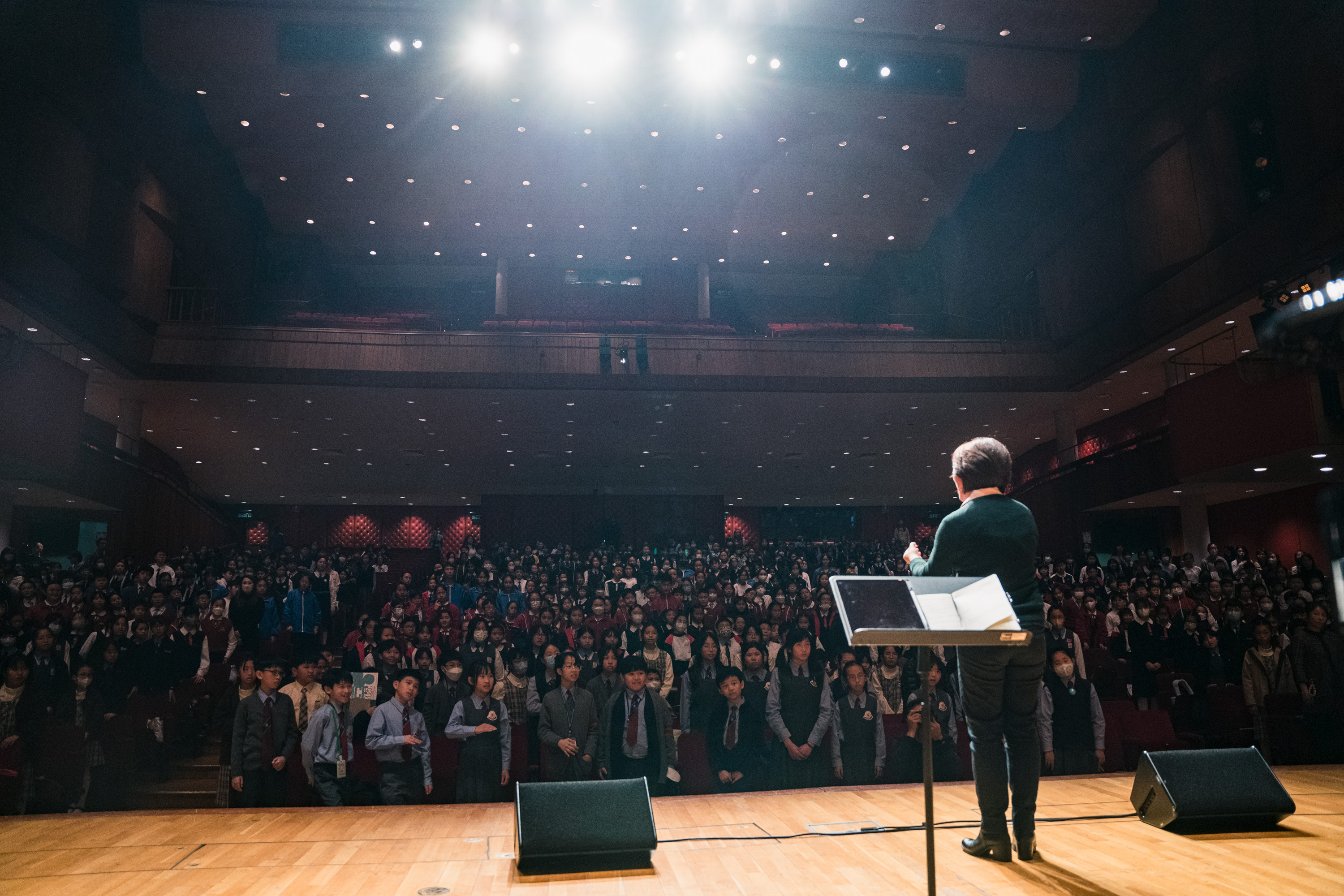 A conductor on stage facing a concert hall filled with schoolchildren