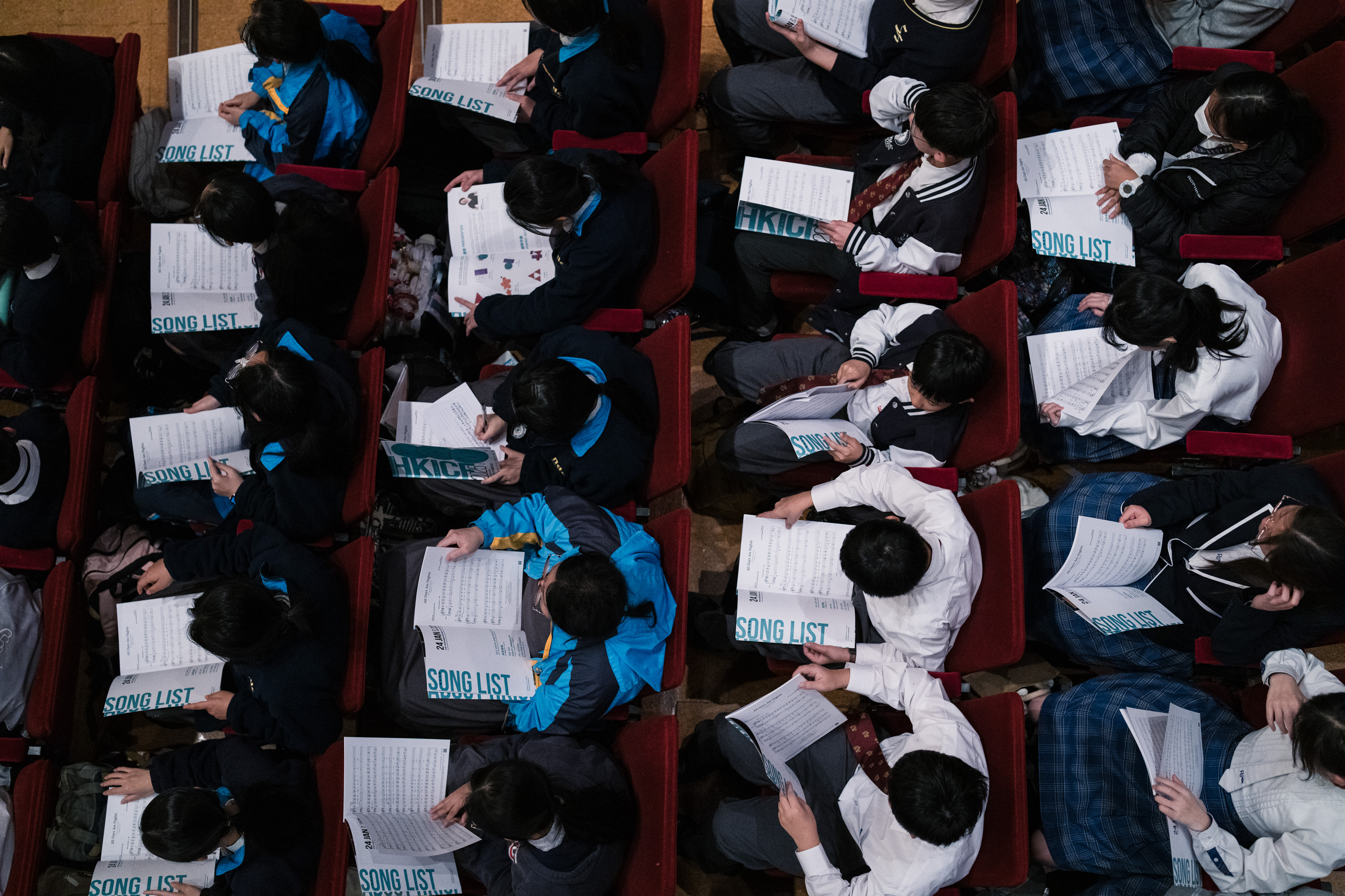 Schoolchildren holding song lists, seen from above in red auditorium seats