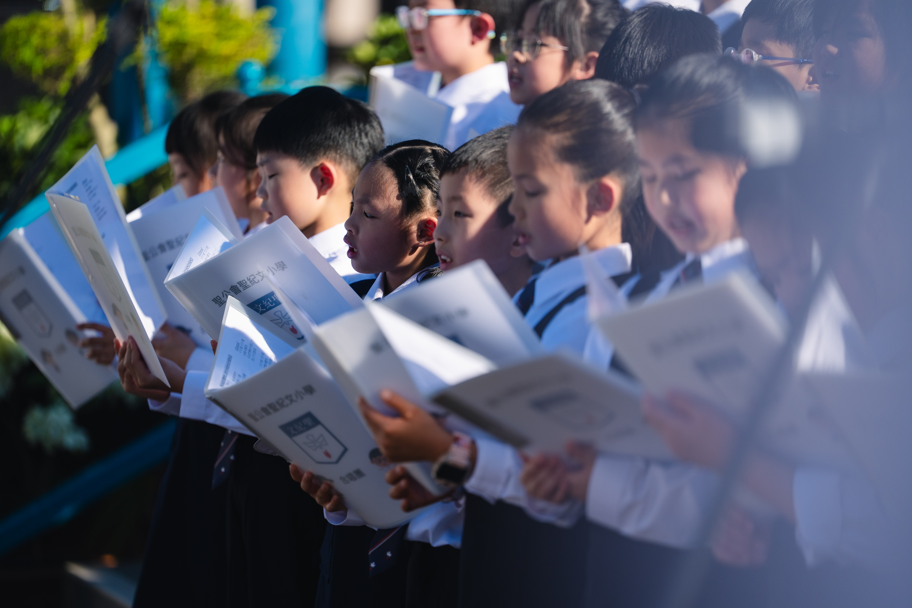 Schoolchildren singing outdoors, each holding a songbook in the sunlight