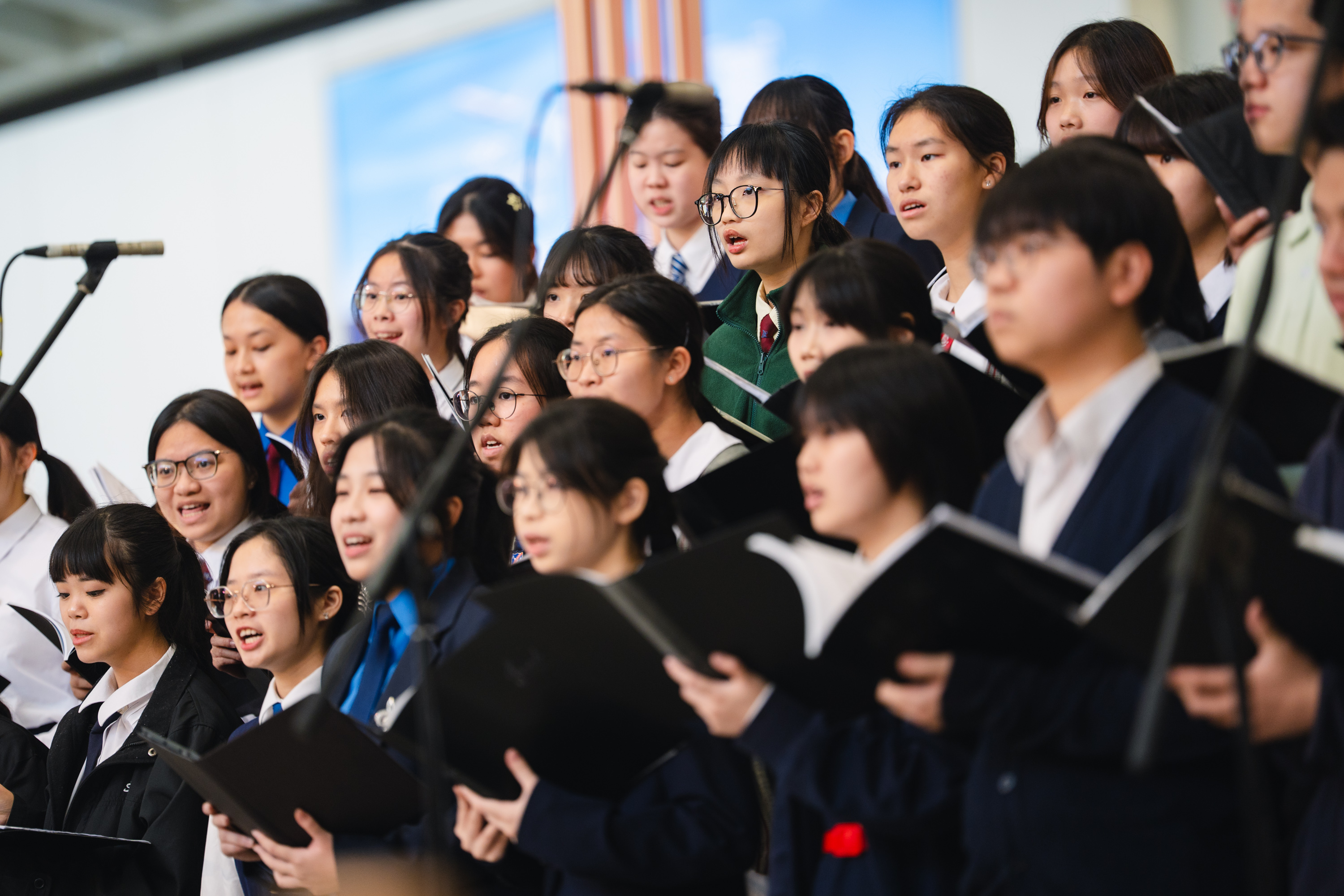 Young singers holding black music folders, singing into microphones at an HKVC programme in December 2025