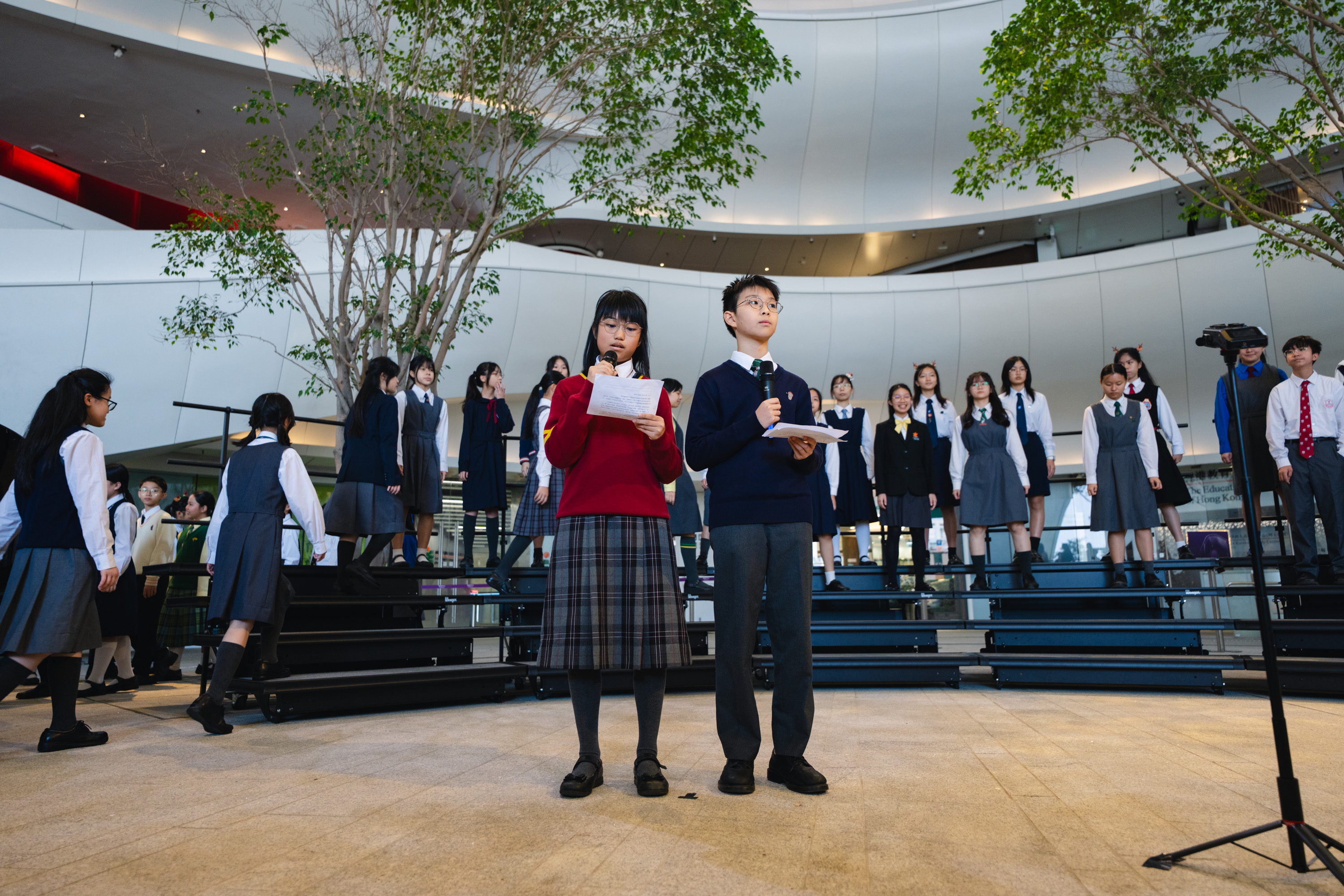 Two young presenters with microphones and scripts introducing a choir performance at an HKVC programme in December 2025
