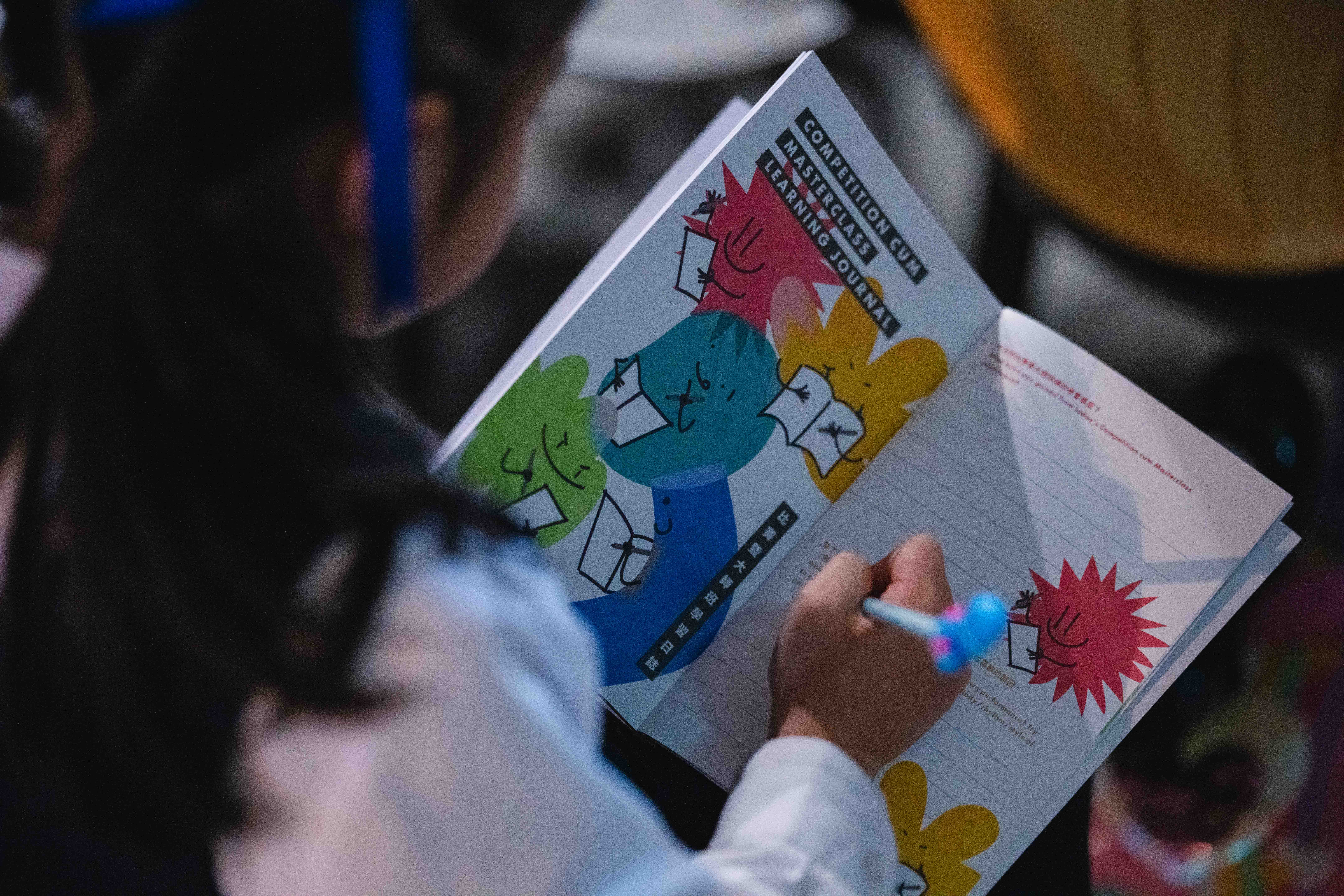 A young participant writing in a colourful illustrated HKVC festival booklet