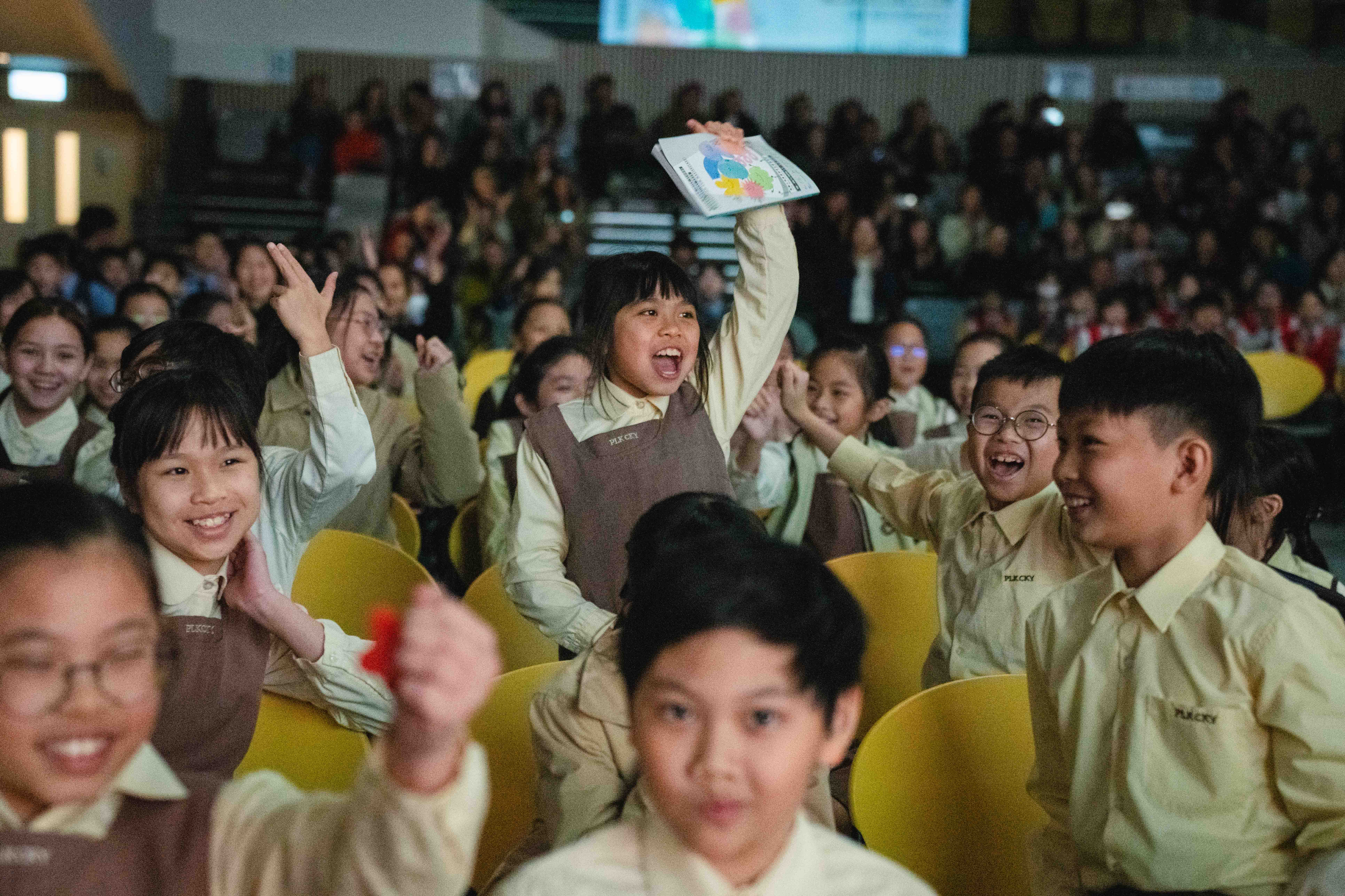 Schoolchildren cheering and waving drawings from yellow auditorium seats