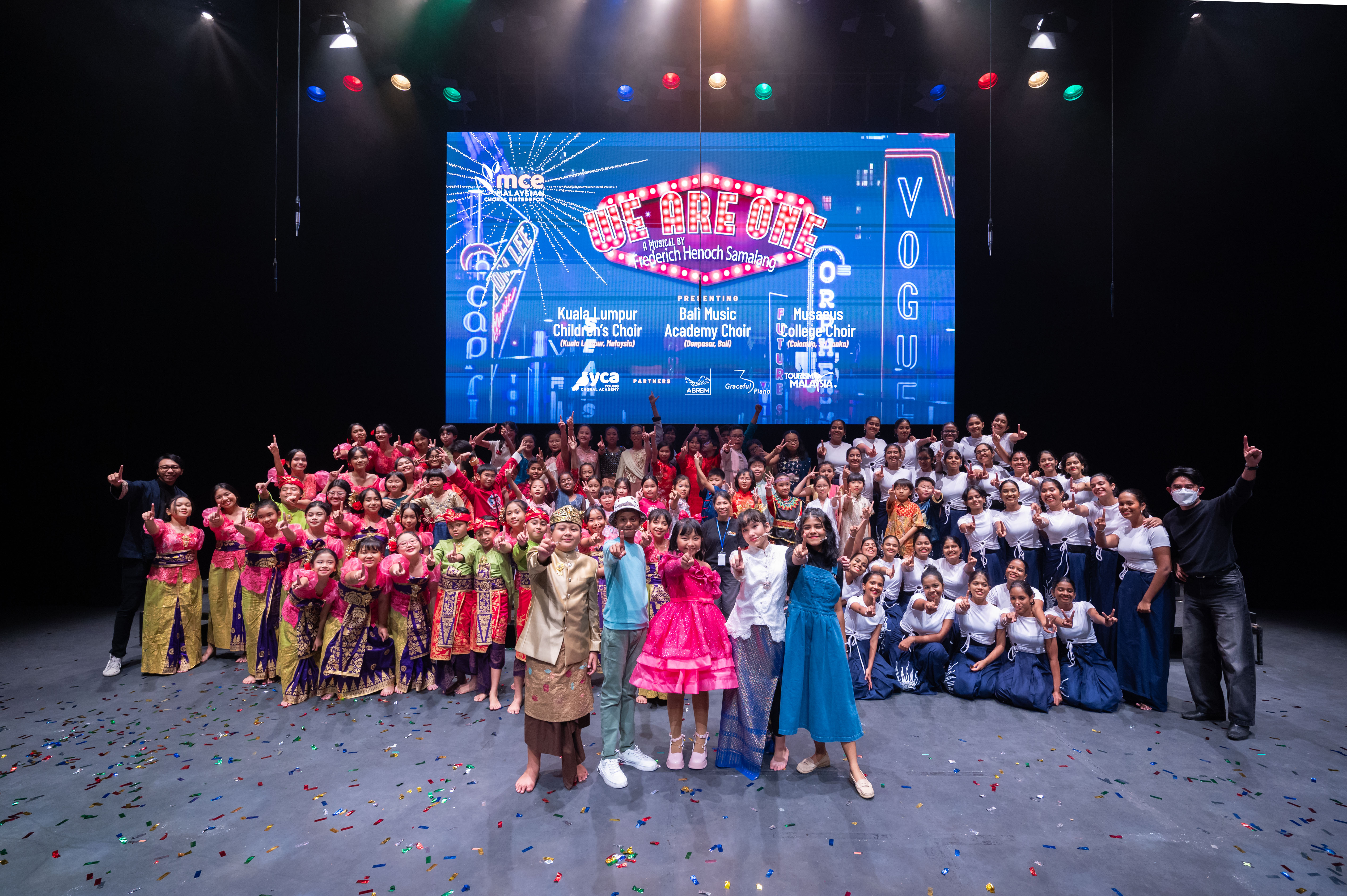 Massed choirs on a confetti-covered stage at the Malaysian Choral Eisteddfod 'We Are One' concert, with participating choirs from Malaysia, Bali and Sri Lanka listed on the LED screen